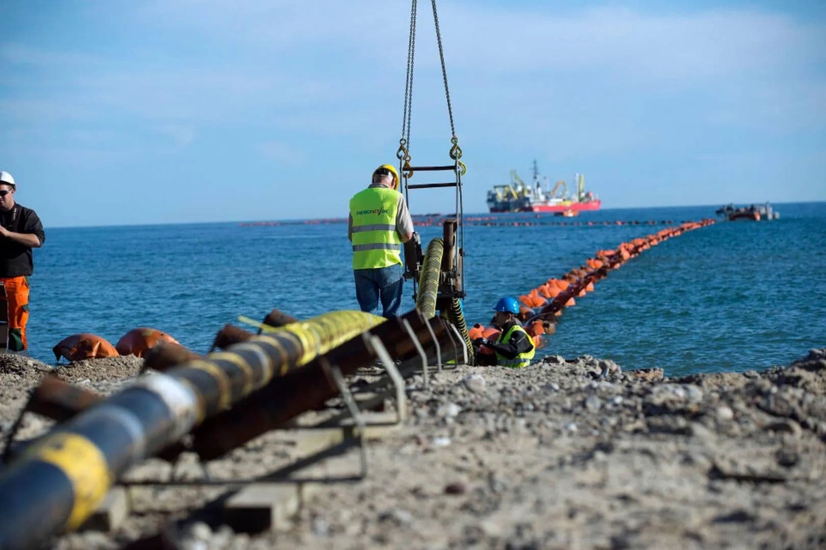 Arbeidere på en steinete kyst legger ut en undersjøisk rørledning i havet, der en lang rekke oransje bøyer strekker seg ut til et fartøy til havs.