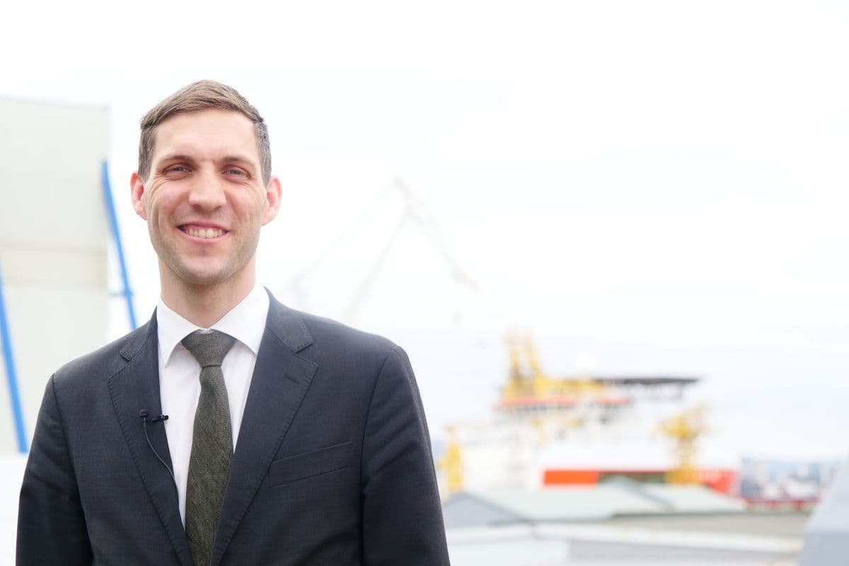 En mann i dress står utendørs og smiler til kameraet, med et industriskip og kraner uskarpe i bakgrunnen.