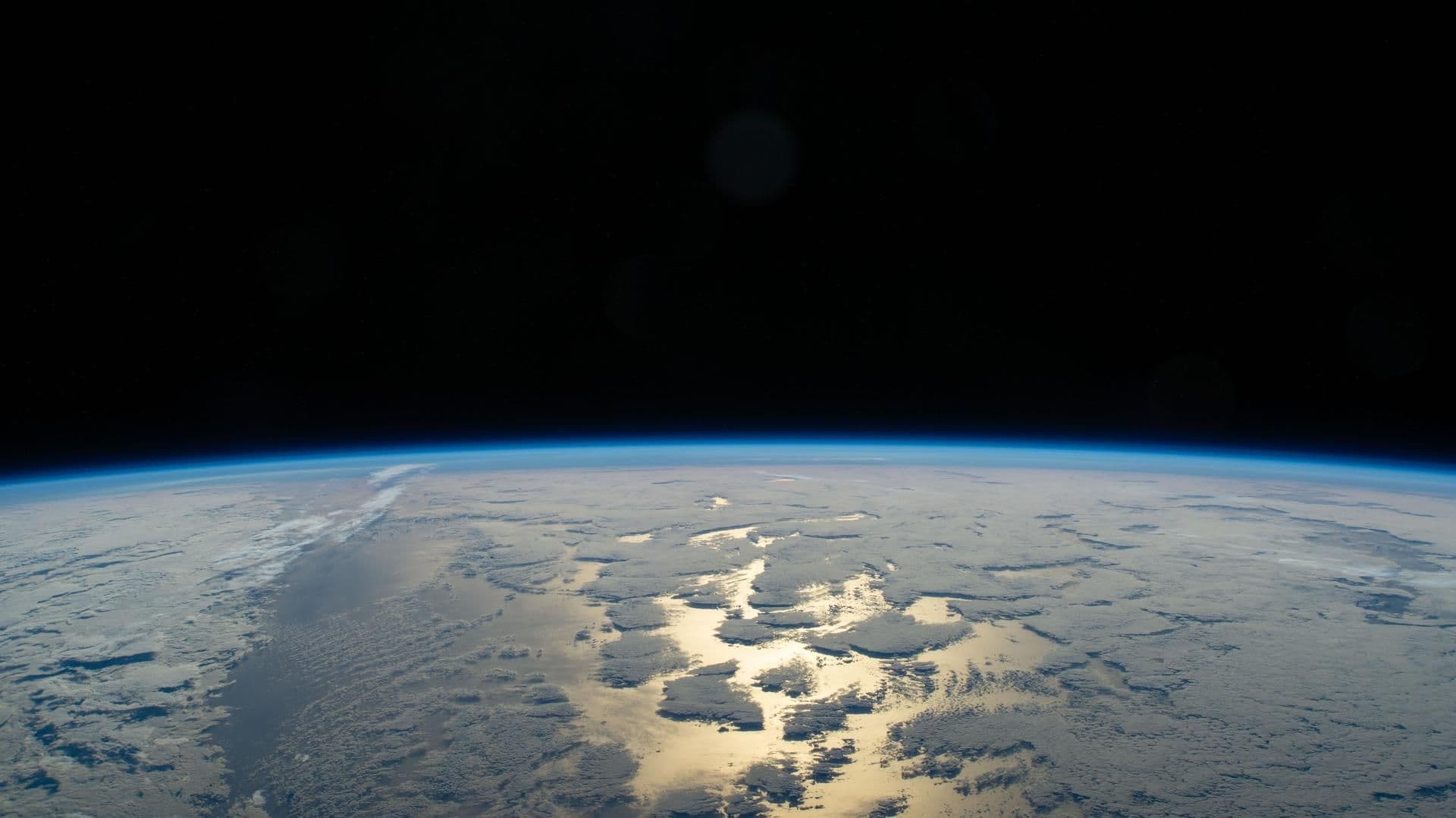 Jordens krumning sett fra verdensrommet, med et blått atmosfærisk skjær, mørk himmel og solbelyste skyer som speiler seg i vannet.