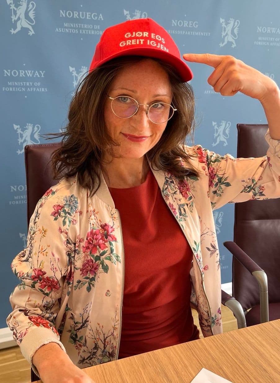 Smilende kvinne med briller og blomstrete jakke som peker på den røde capsen sin med teksten «GJØR EG GREIT IGJEN».