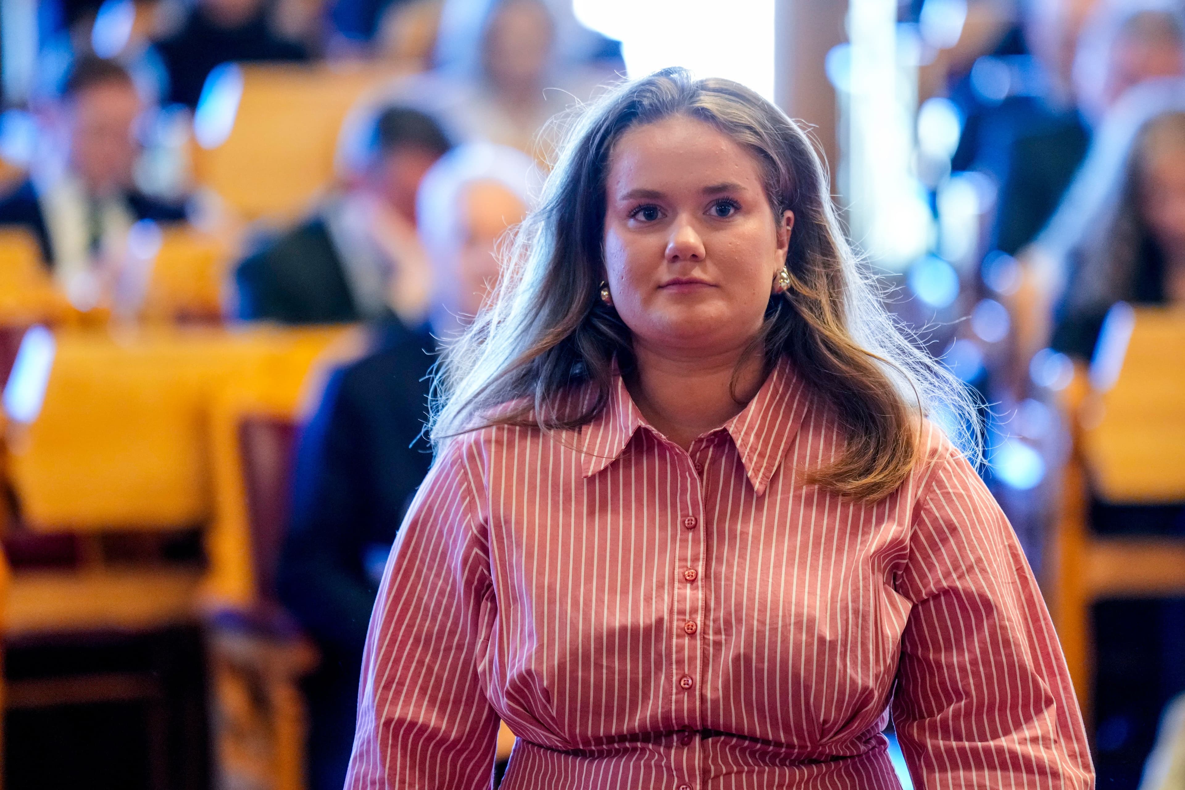 En ung kvinne med langt, lysebrunt hår, perleøredobber og en rosa- og hvitstripet skjorte ser rett mot betrakteren i formelle omgivelser med uskarp bakgrunn.