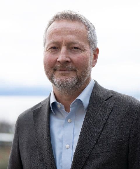 Portrett av en smilende mann med grått hår og skjegg, iført blå skjorte og mørk dressjakke.