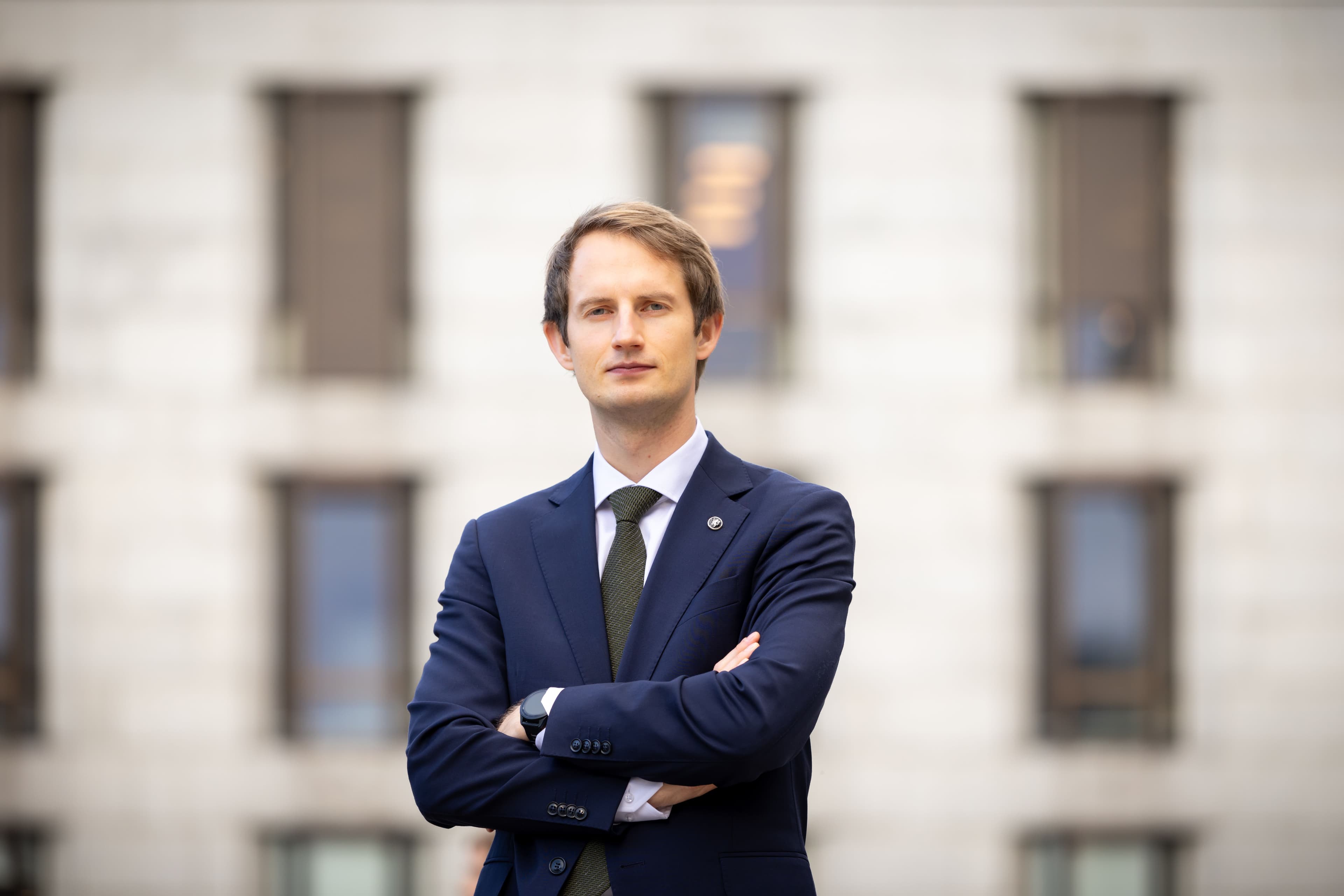 Bildet viser Aleksander Stokkbø (H) i dress og slips som står med korslagte armer foran en uklar bygning.