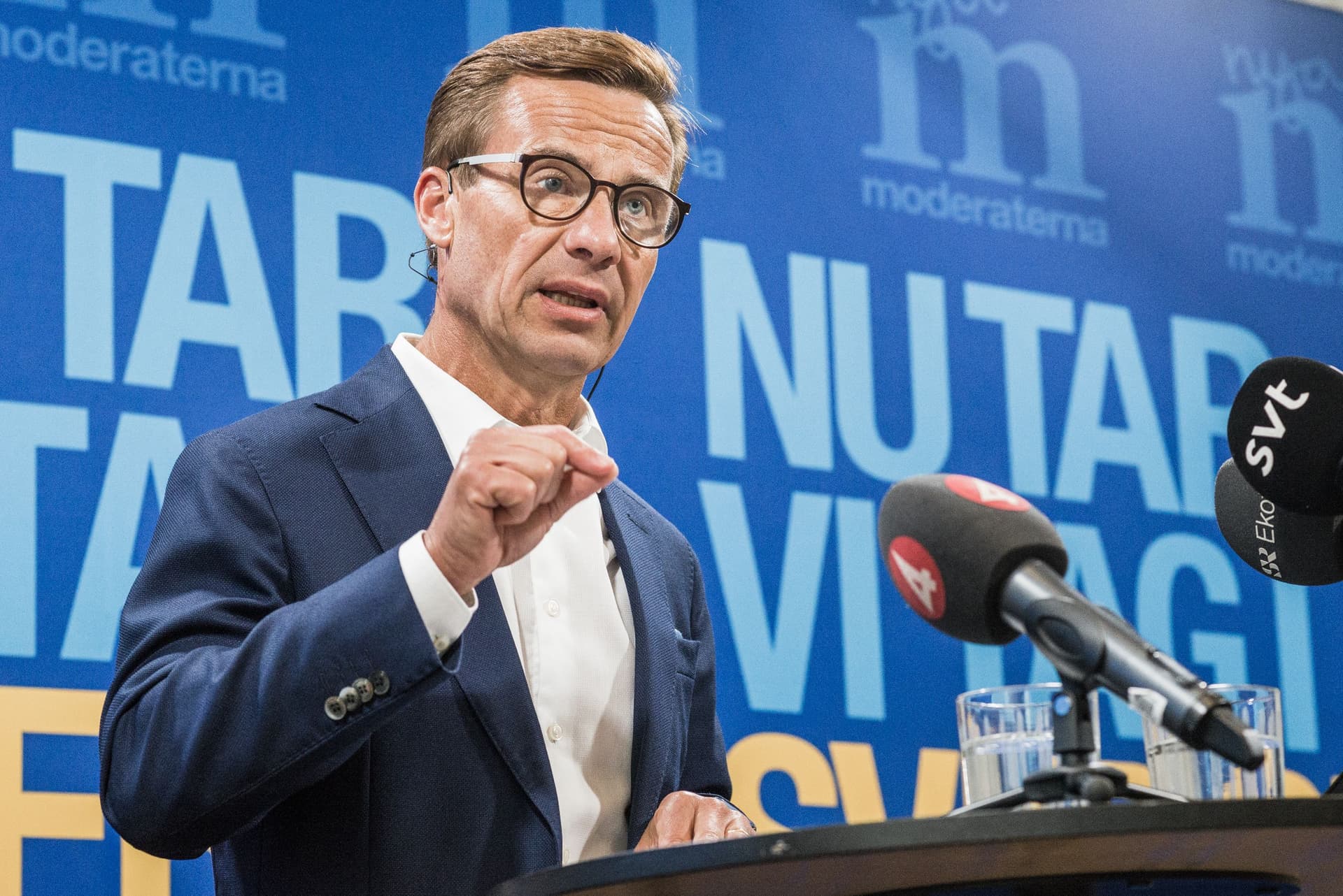 Sveriges statsminister Ulf Kristersson taler på en pressekonferanse og gestikulerer med en knyttet neve, foran en blå bakgrunn med partilogoen og teksten «moderaterna», med mikrofoner i forgrunnen.