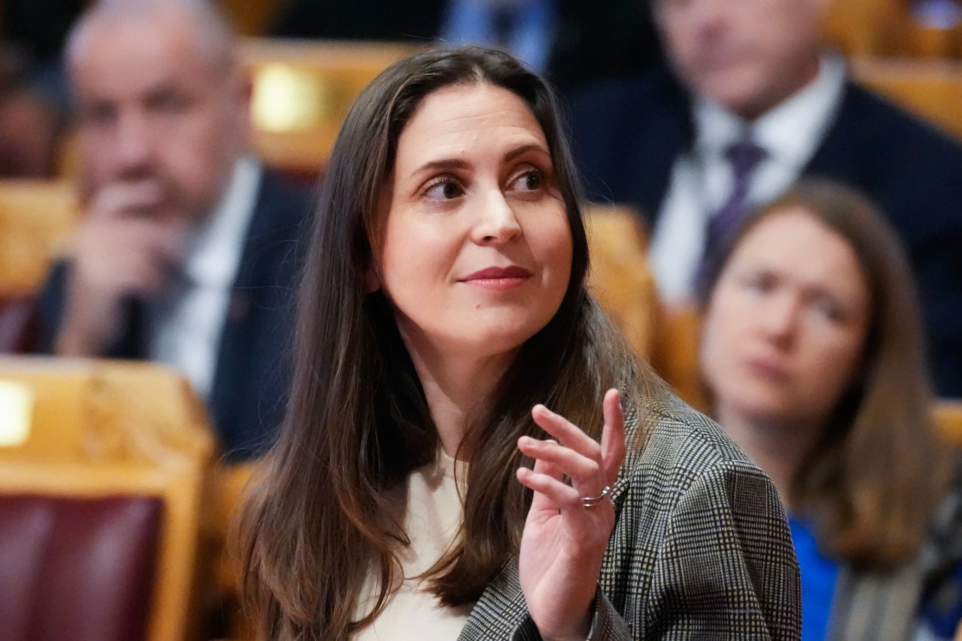 En kvinne med langt mørkt hår, iført en rutete blazer, ser mot høyre og gestikulerer med venstre hånd i et uskarpt møterom.