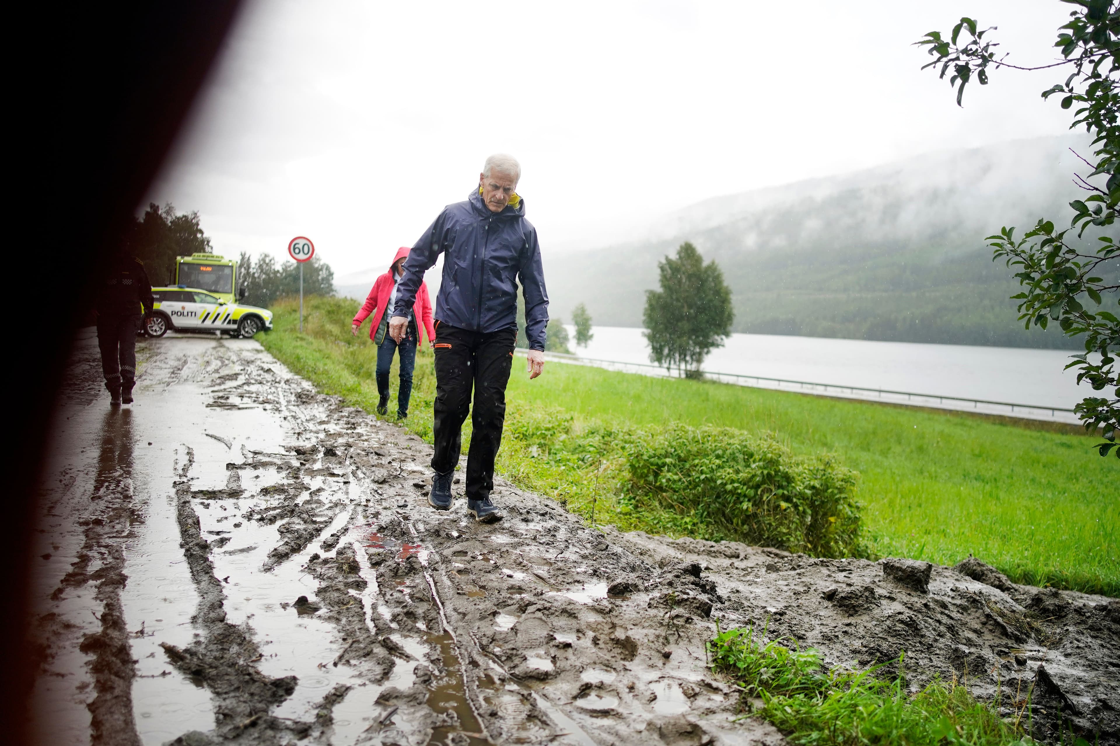 Jonas Gahr Støre går på en svært gjørmete vei, forbi en politibil og andre mennesker, med en innsjø og tåkete åser i bakgrunnen.