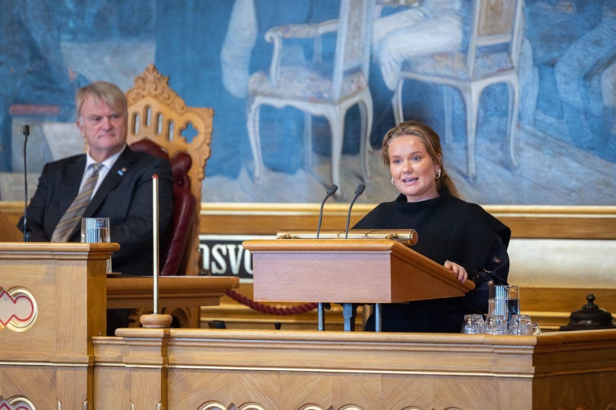 En kvinne taler på et podium i en formell sal, mens en mann sitter ved siden av henne på en trestol. I bakgrunnen skimtes et veggmaleri og tomme stoler.