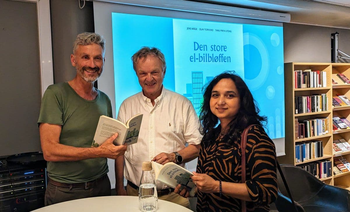 Tre personer står sammen innendørs og holder kopier av en bok med tittelen «Den store el-bilbløffen». De ser ut til å være i et rom med bokhyller og en skjerm i bakgrunnen som viser bokomslaget.