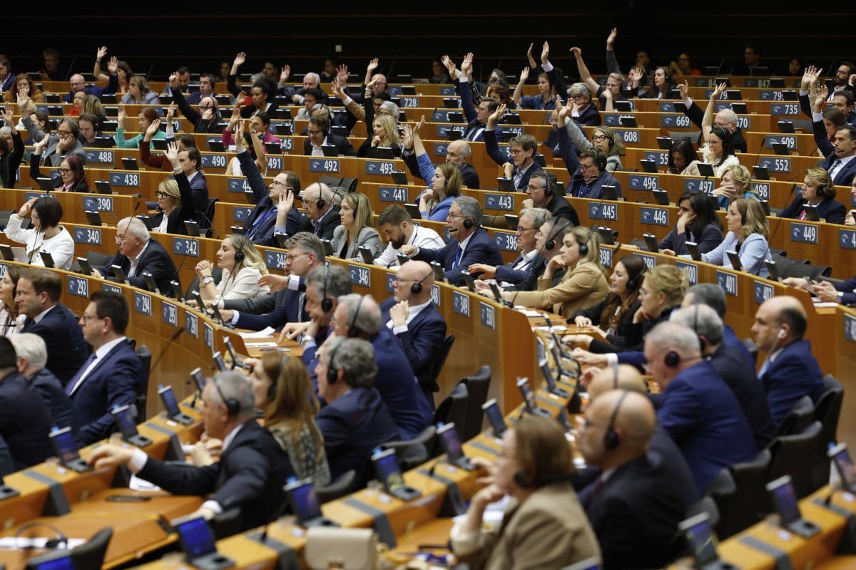 En stor gruppe mennesker som sitter i et auditorium, noen rekker opp hånda, har nummererte pulter og bruker hodetelefoner, noe som tyder på et formelt møte eller en forsamling.