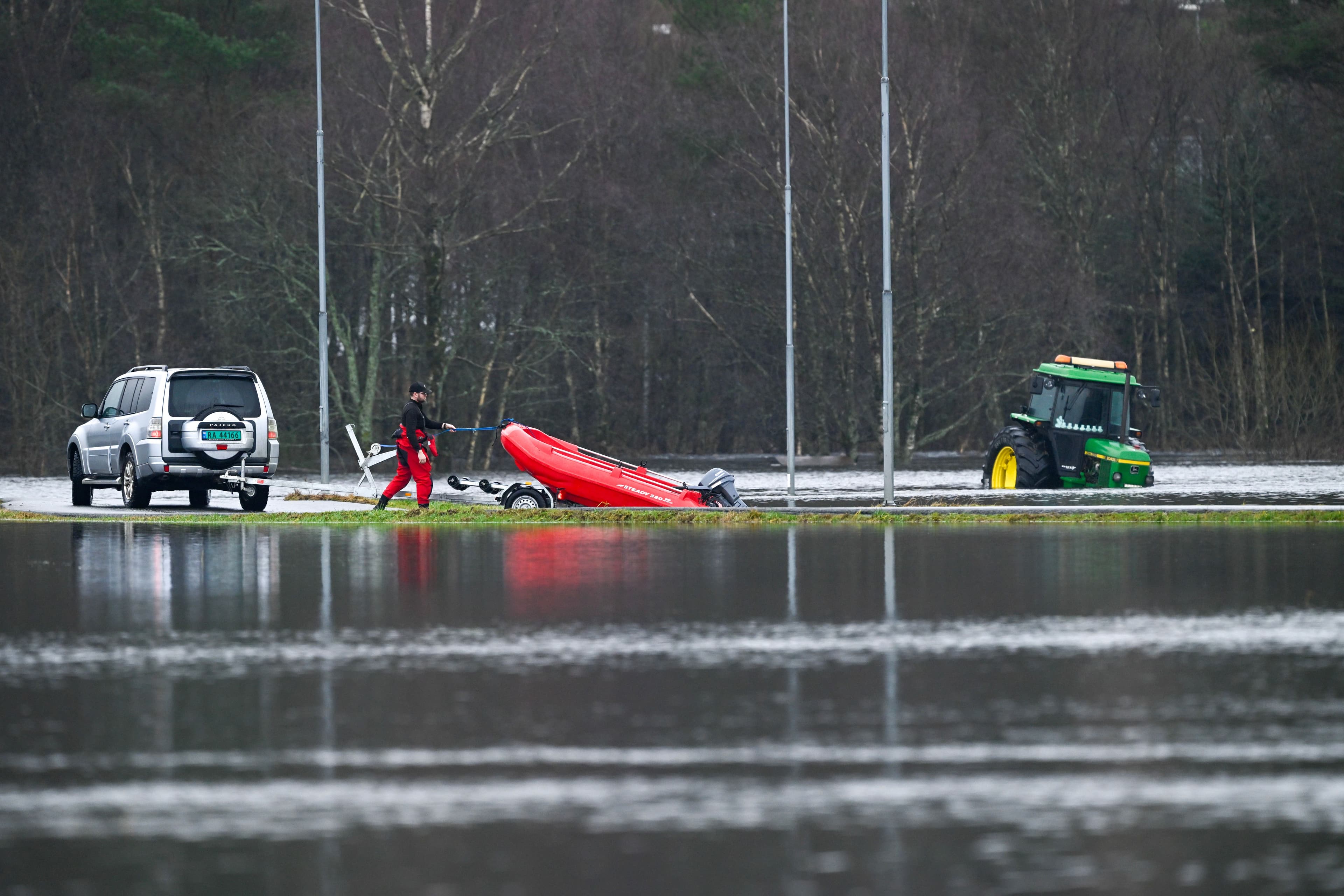 Oversvømt landskap med en person som drar en rød oppblåsbar båt, en SUV og en delvis nedsunket grønn traktor.