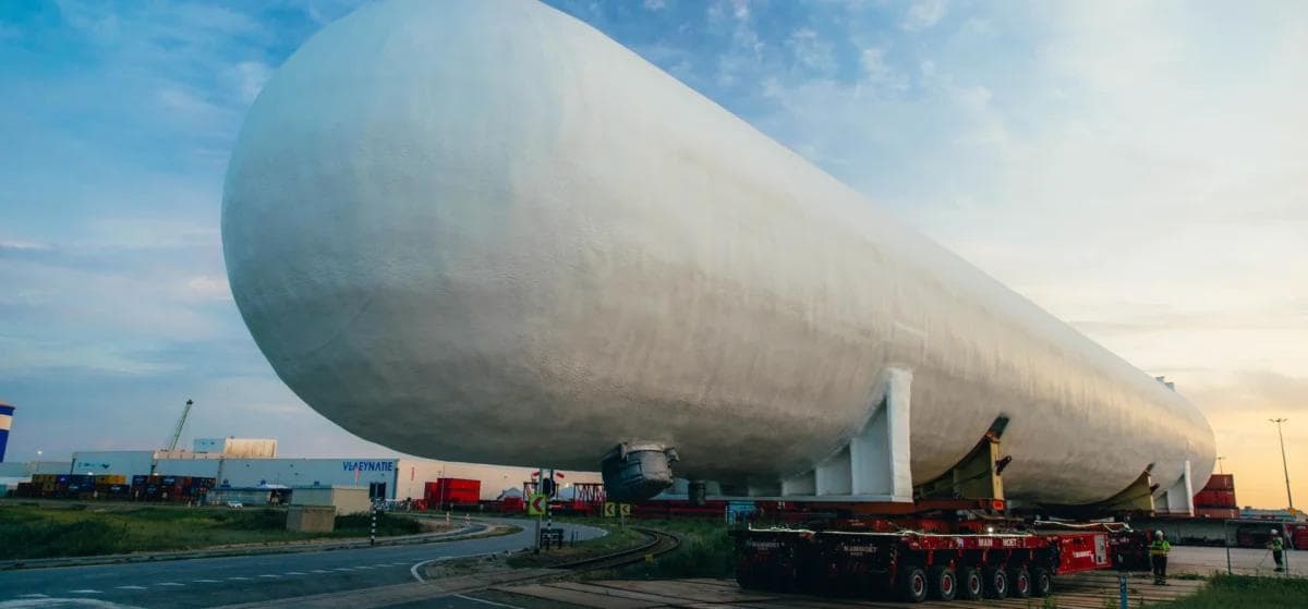 En stor, sylinderformet industritank transporteres på et kjøretøy med flere hjul langs en vei i nærheten av lagerbygninger og kraner i solnedgangen.