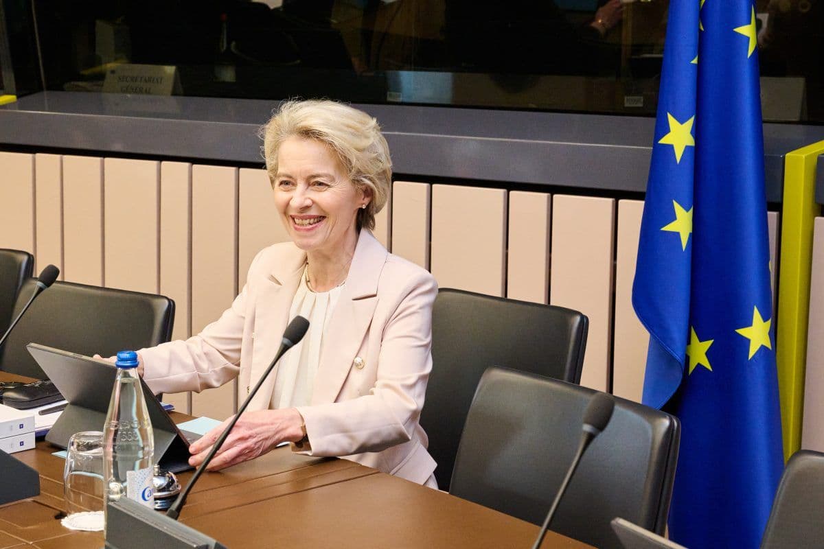 En person som sitter ved et konferansebord med mikrofoner og en bærbar datamaskin, nær et EU-flagg.