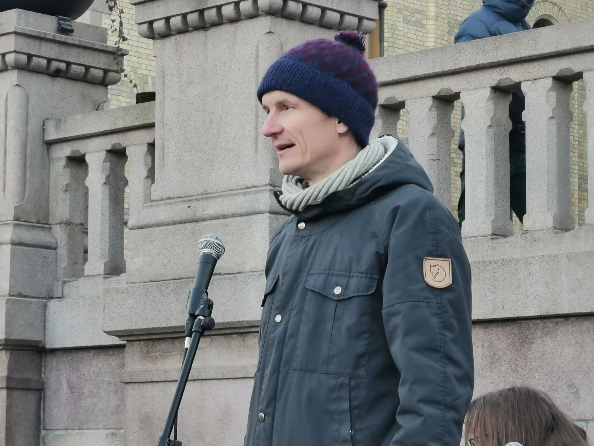 En mann i blå jakke og lue snakker i en mikrofon utendørs.