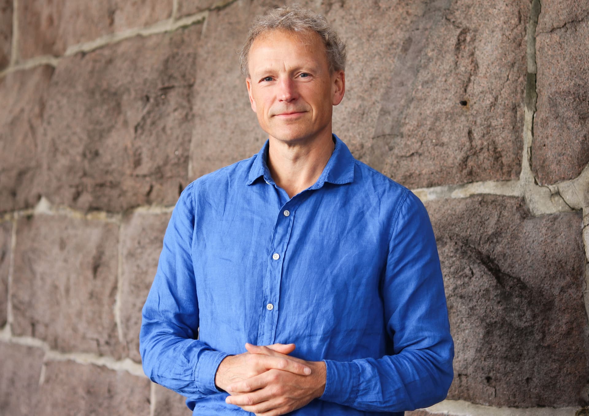 En mann med gråsprengt hår i blå skjorte står og smiler med hendene foldet foran seg mot en steinmur.