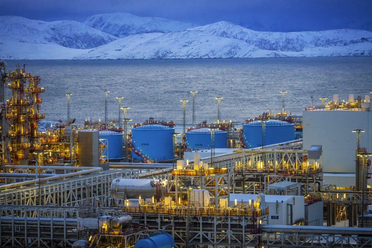 Industrielt olje- eller gassbehandlingsanlegg med store lagertanker og komplekse rørledninger, med snødekte fjell og vann som bakgrunn.