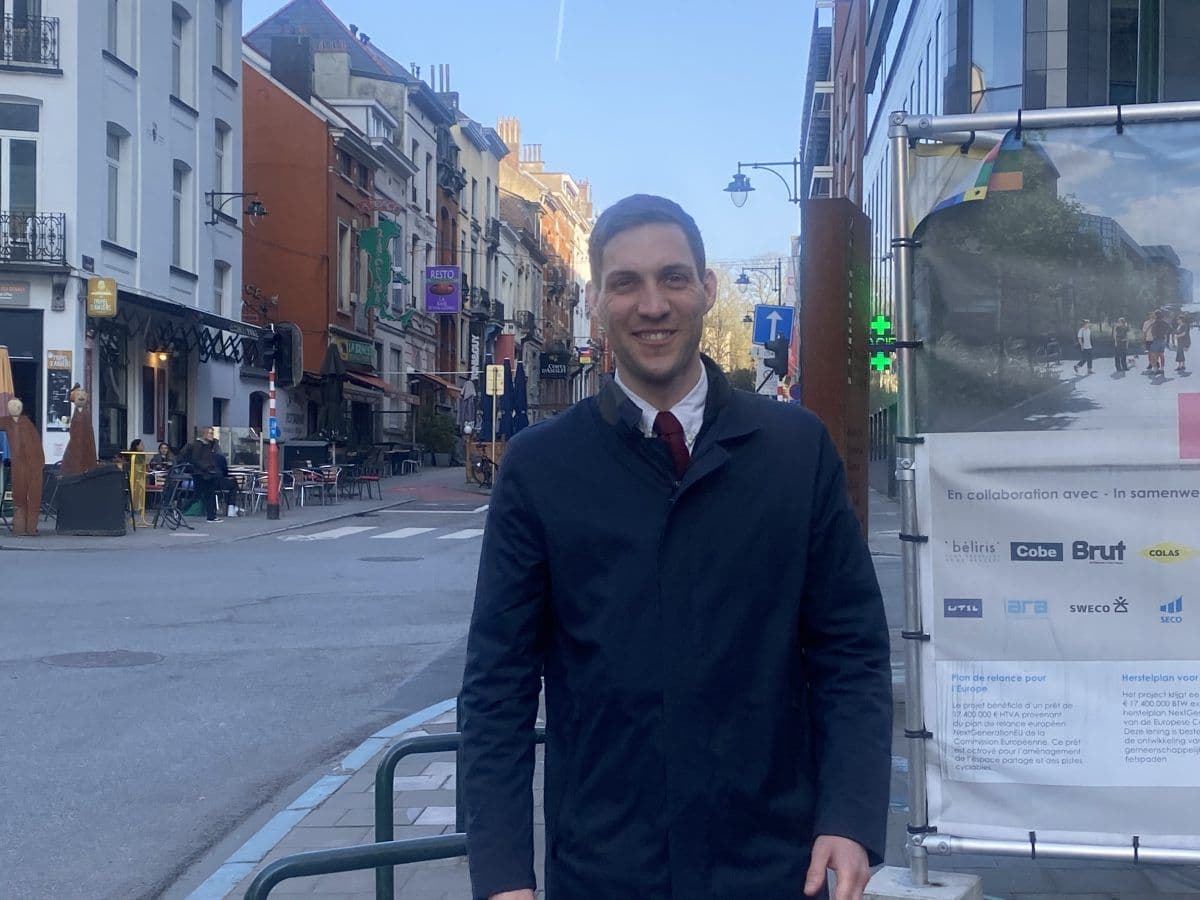 Smilende person i mørk frakk står i en bygate, med bygninger og parkerte sykler i bakgrunnen.