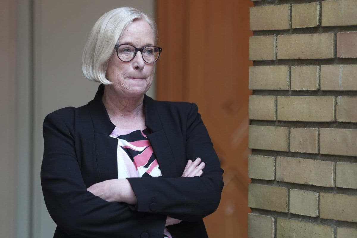 En kvinne med kort blondt hår og briller står med armene i kors, iført en svart blazer og mønstret topp, ved siden av en murvegg.