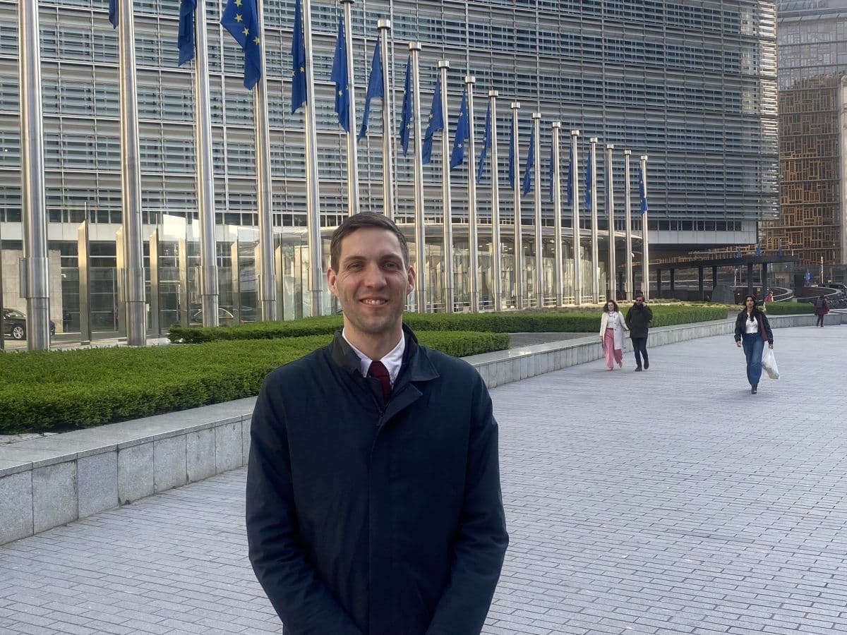 En person står og smiler foran en moderne bygning med flere EU-flagg på flaggstenger. Andre mennesker går i bakgrunnen.