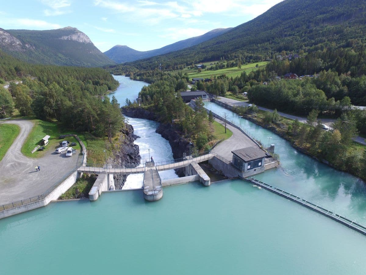 Luftfoto av en turkis elv som renner gjennom en demning omgitt av trær og fjell, med en vei og kjøretøy på venstre side.