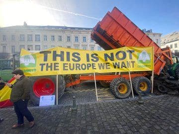 Protestscene med traktorer og banner hvor det står «Dette er ikke Europa vi vil ha» på et brosteinstorg, med folk i nærheten.