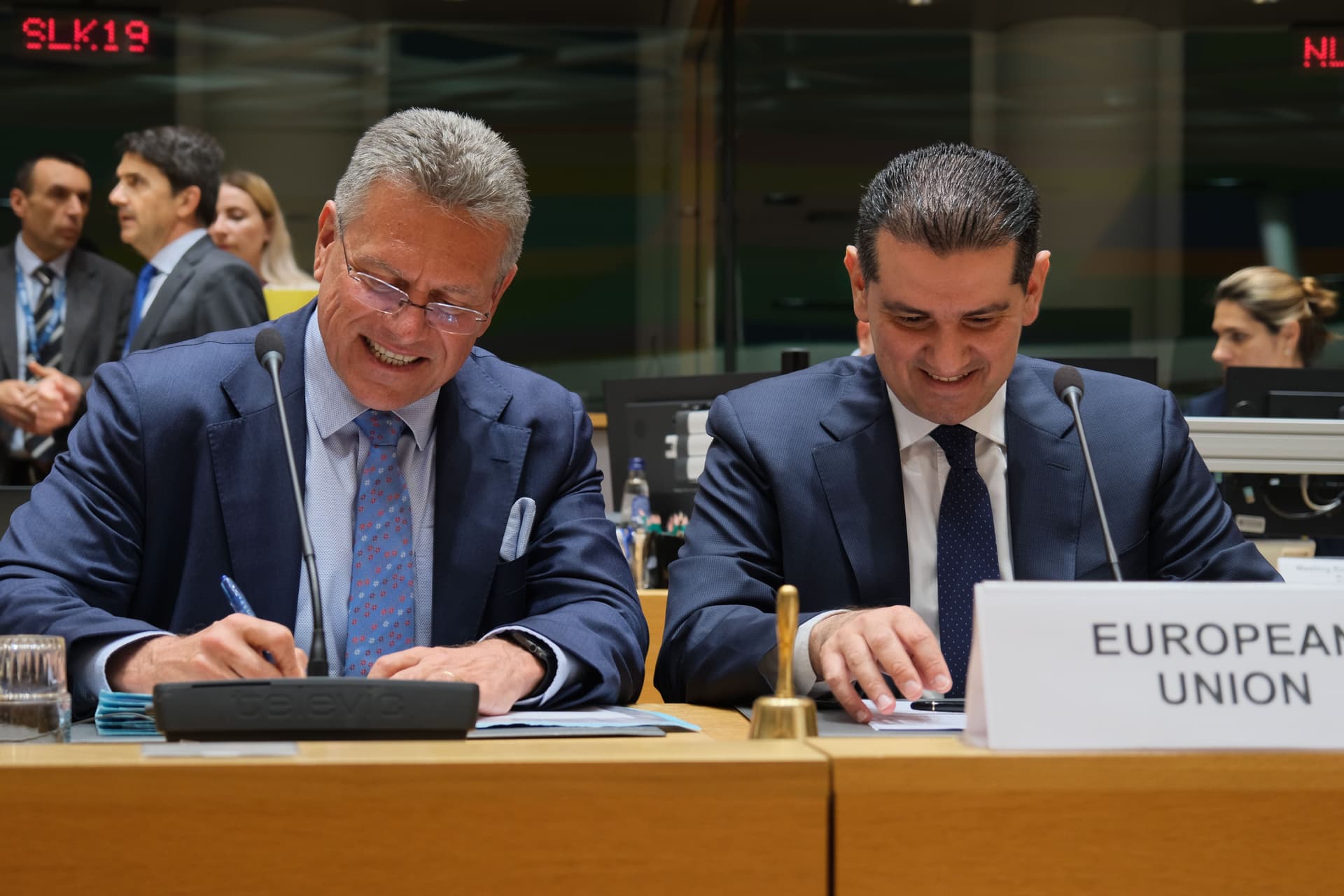 Two men in suits at a European Union meeting, one signing intently, the other smiling.