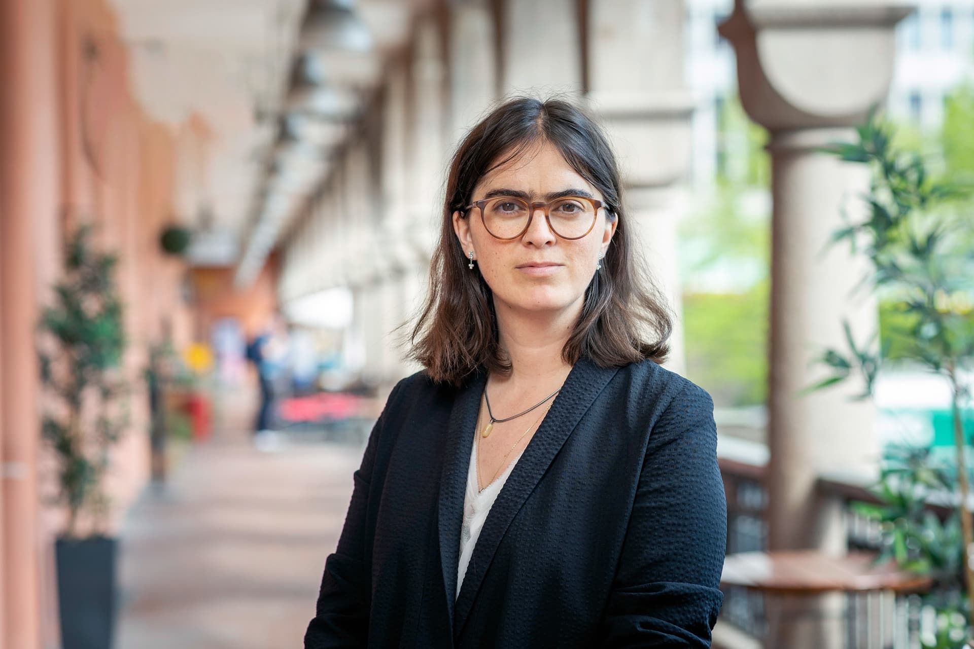 En kvinne med mørkt hår og briller, iført en mørk blazer, ser rett inn i kameraet under en overbygd gangvei.