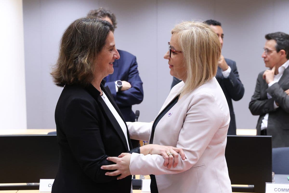 To kvinner i forretningsantrekk håndhilser og smiler til hverandre i et konferanserom med andre mennesker i bakgrunnen.