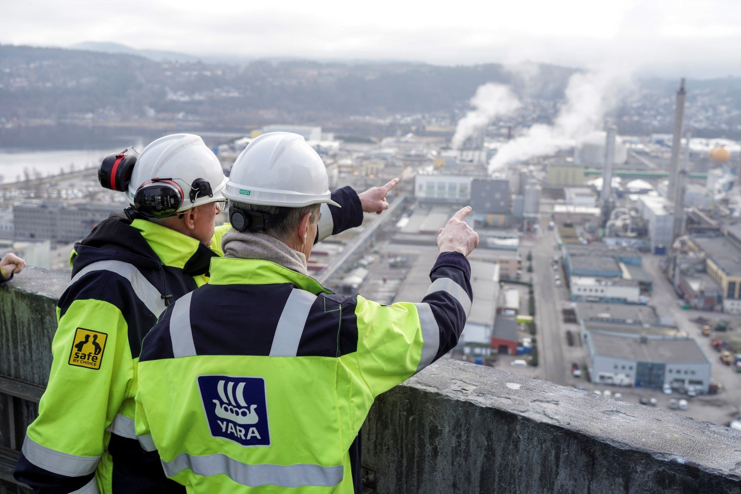 Jonas Gahr Støre og en annen mann, begge med ryggen til, peker mot industrianlegg.