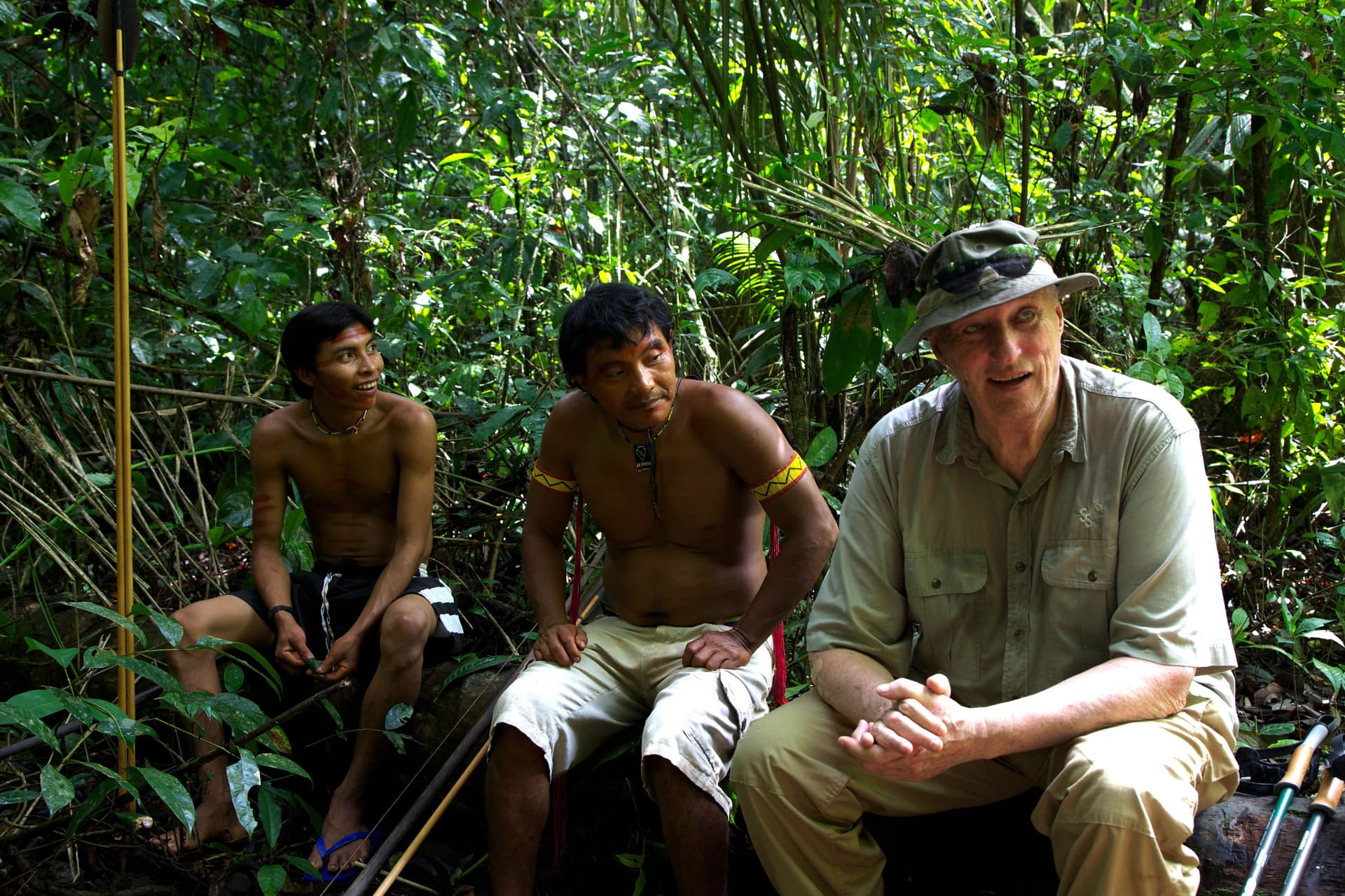 An older white man and two shirtless Indigenous men sit together in a lush jungle.