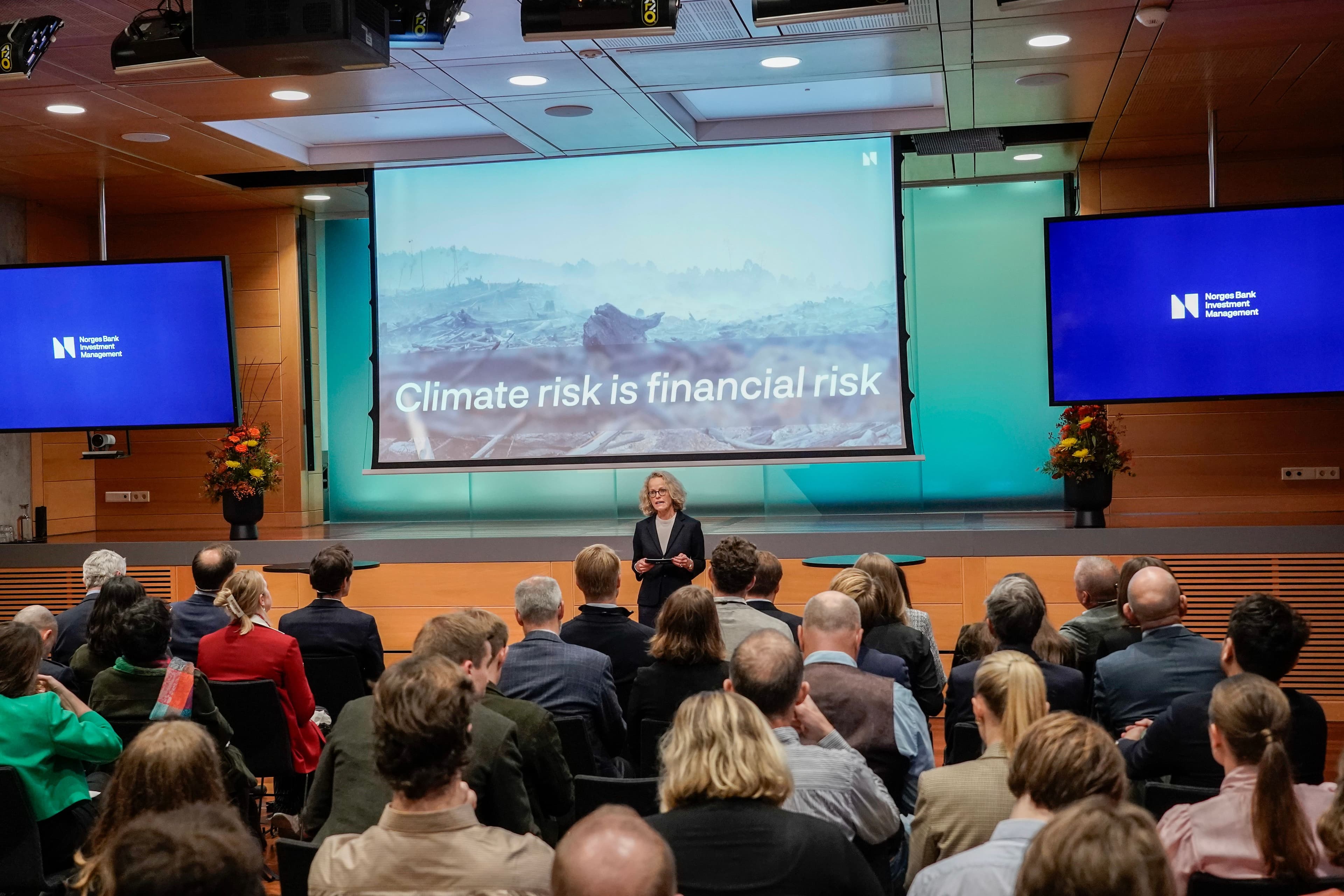 Kvinne på scene presenterer klimahandlingsplan foran publikum, med stor skjerm bak som viser grafikk om klimarisiko og investeringer.