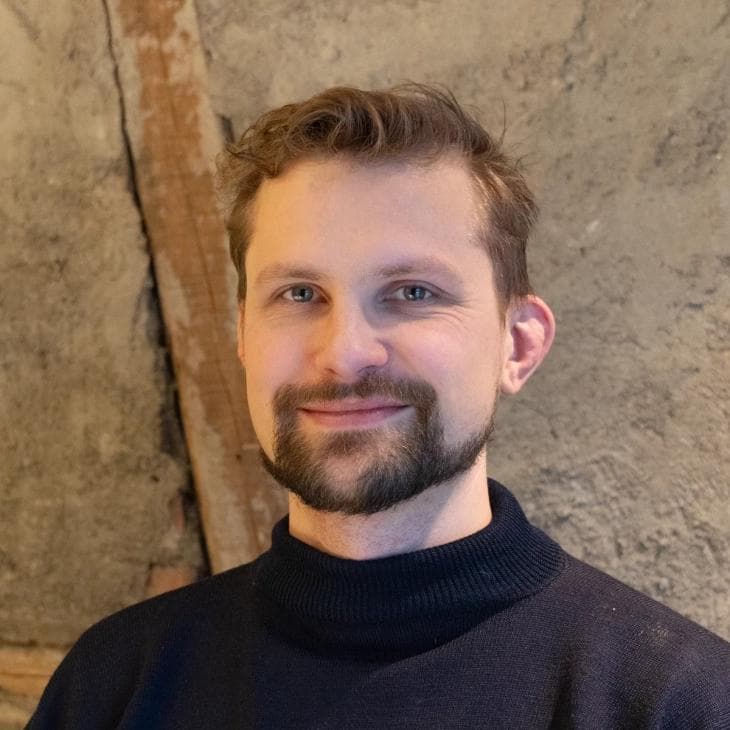 En smilende mann med lysebrunt hår og fippskjegg har på seg en mørk pologenser foran en teksturert vegg med en trebjelke.