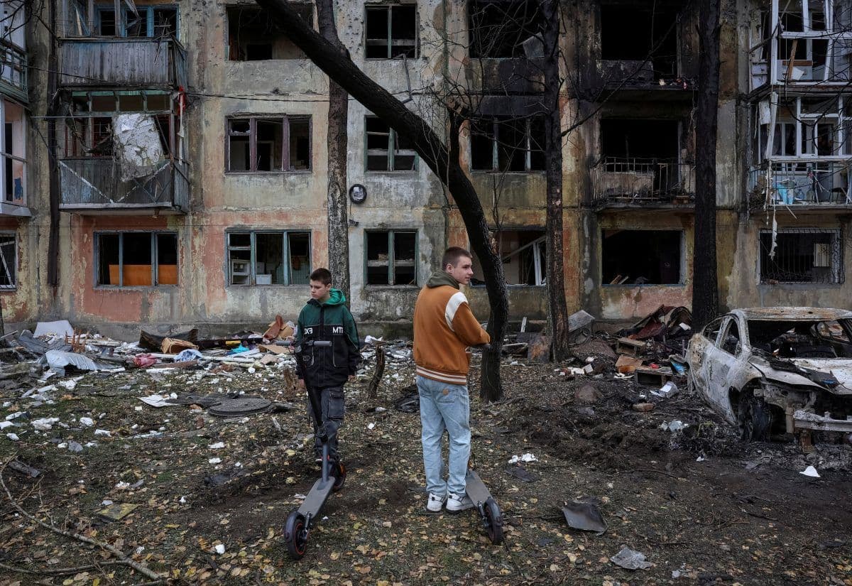 To gutter på elsparkesykler står foran en utbrent boligblokk med store skader, og rundt omkring ligger vrakrester og en ødelagt bil.