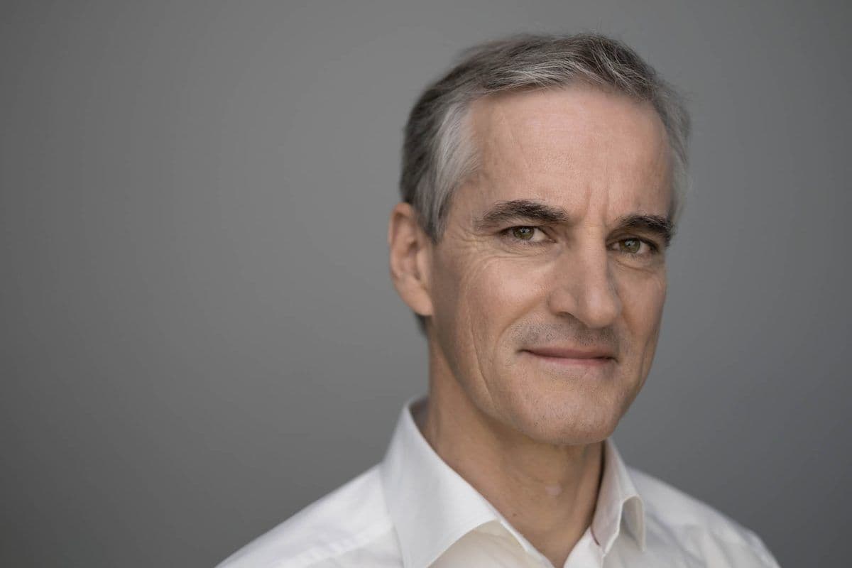 En middelaldrende mann med grått hår og hvit skjorte med krage ser inn i kameraet og smiler lett mot en grå bakgrunn.