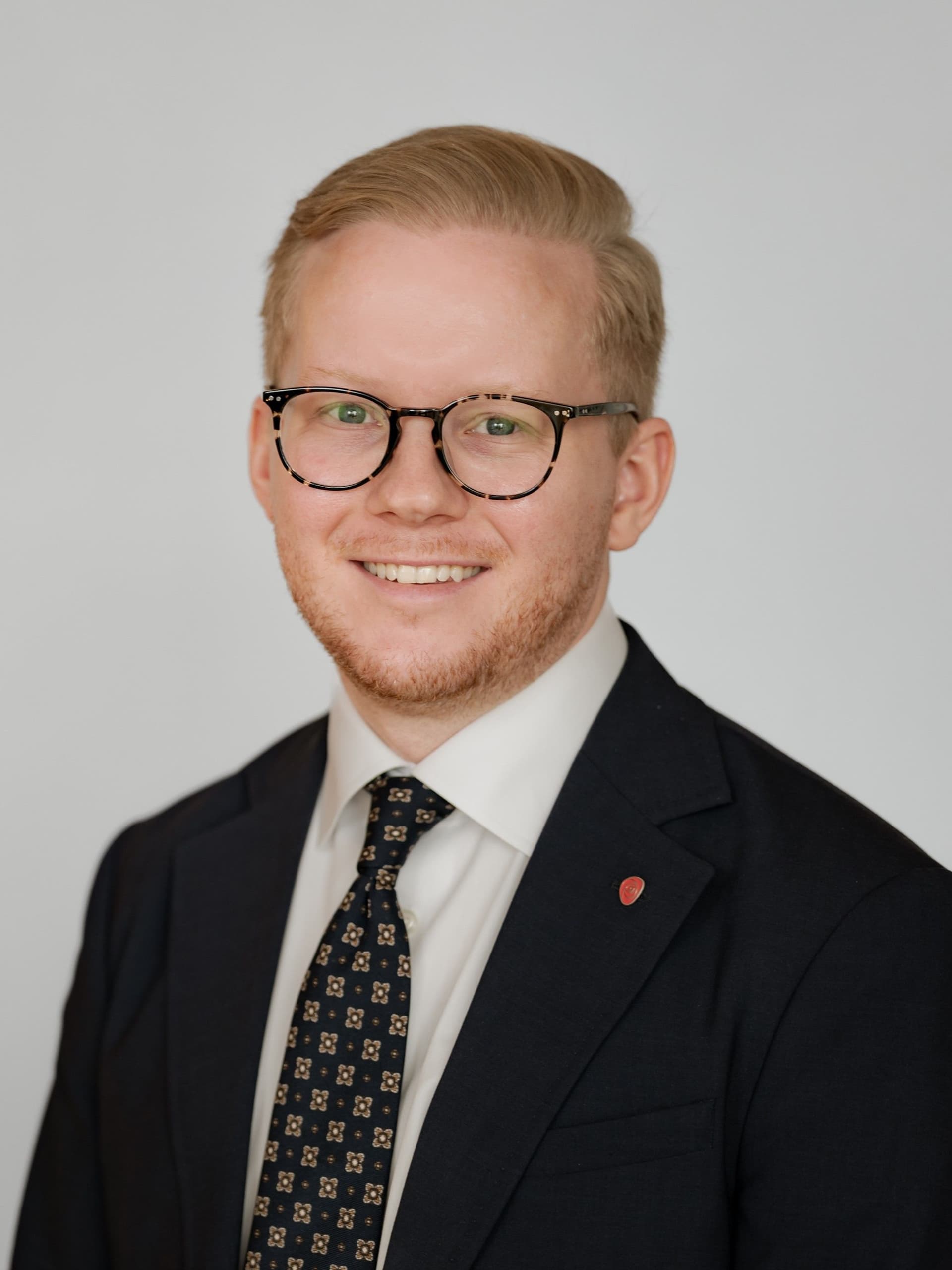 En smilende mann med blondt hår, briller og lett skjegg har på seg en mørk dress, mønstret slips og en rød jakkeslagsnål.