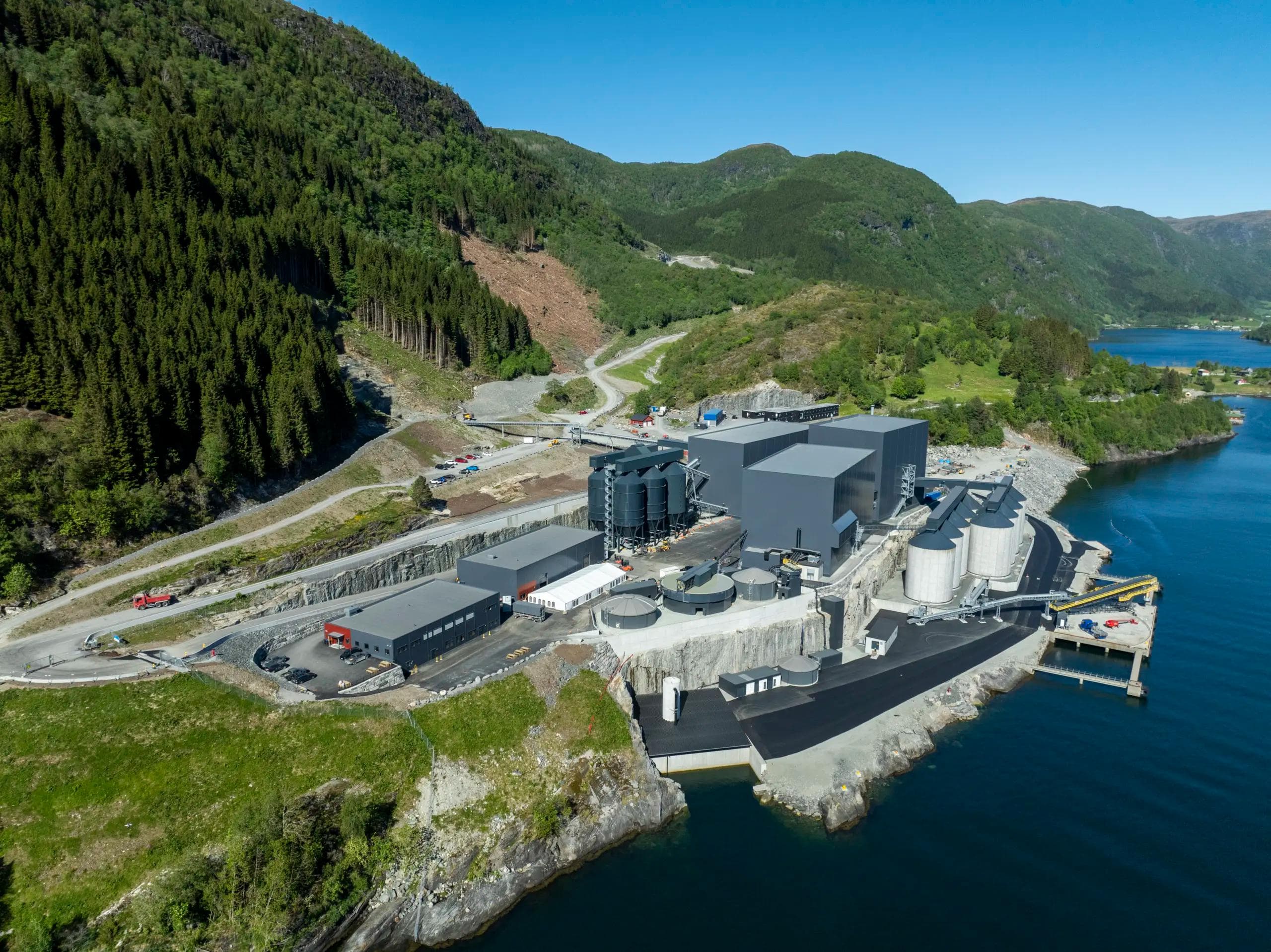 Flyfoto av et moderne industrikompleks med grå bygninger og siloer, plassert ved en fjord og omgitt av skogkledde fjell.