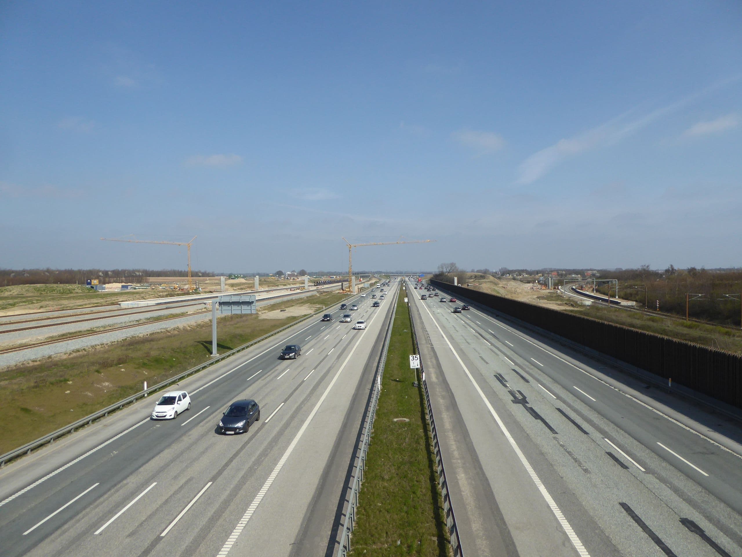 Flerfelts motorvei med lett trafikk, med jernbanebygging på venstre side og støyskjerm på høyre side, under blå himmel.