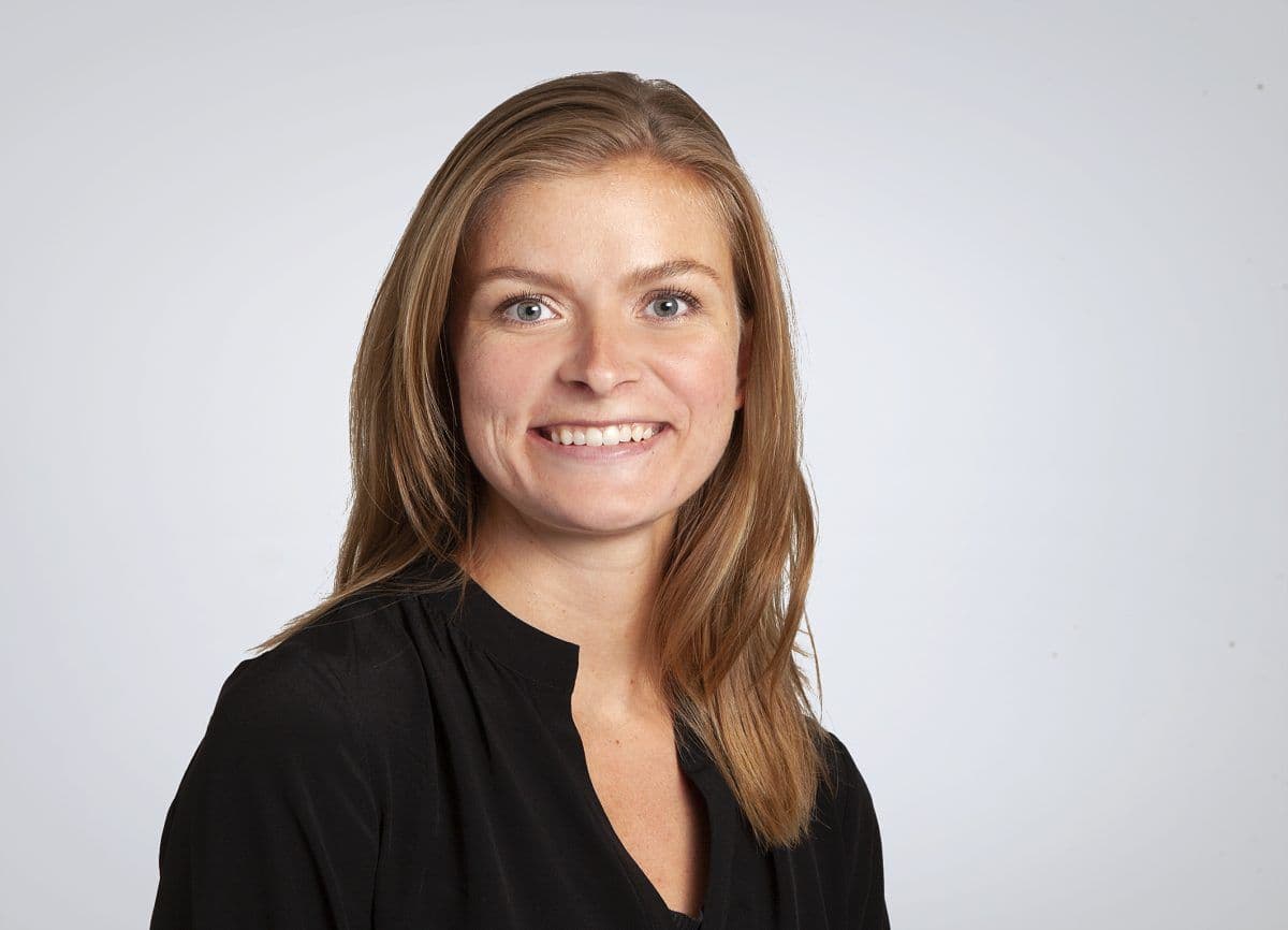 En kvinne med langt lysebrunt hår, iført en svart topp, smiler til kameraet mot en vanlig lysegrå bakgrunn.