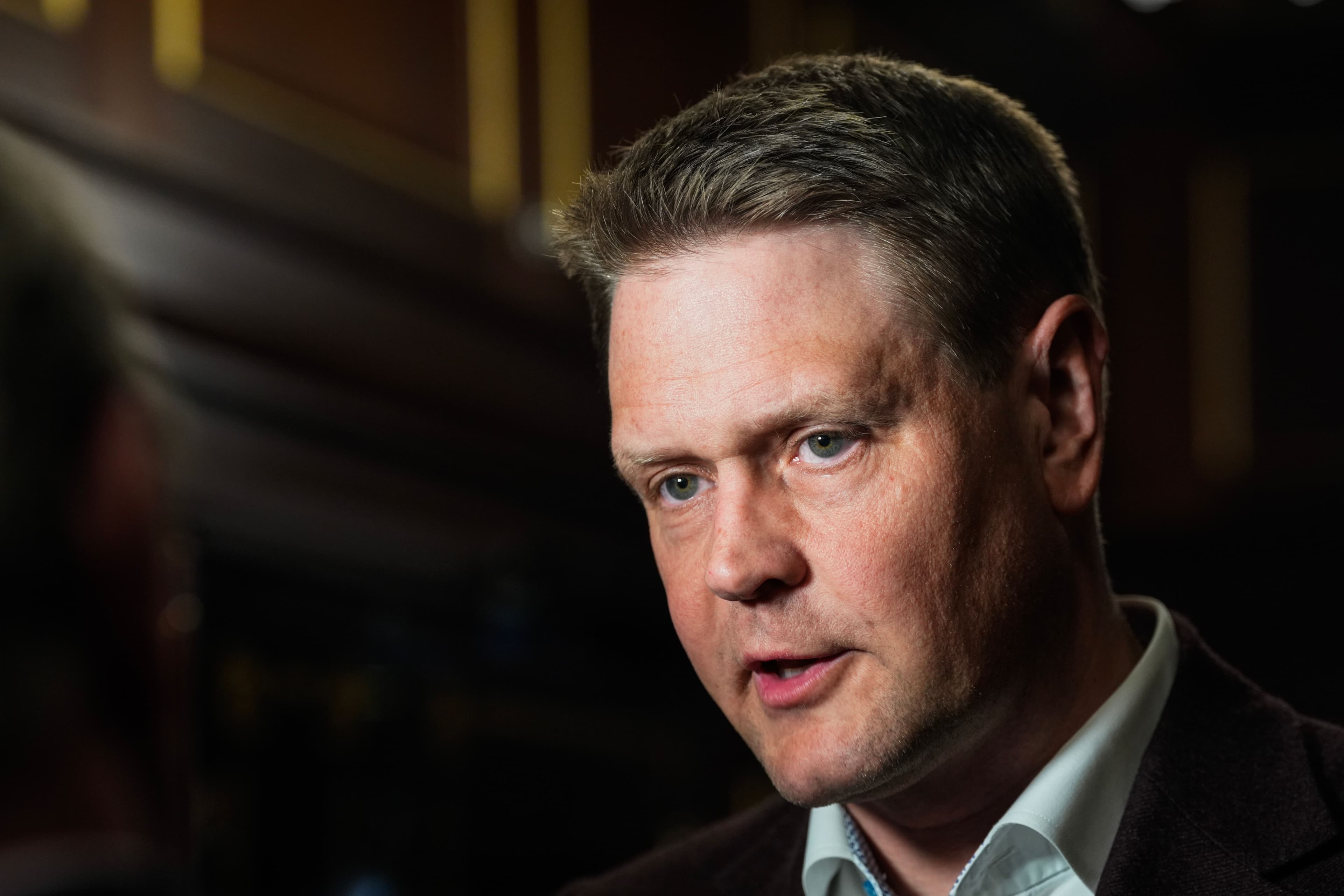 En lyshudet mann med lysebrunt hår og blå øyne, iført en brun jakke, snakker.