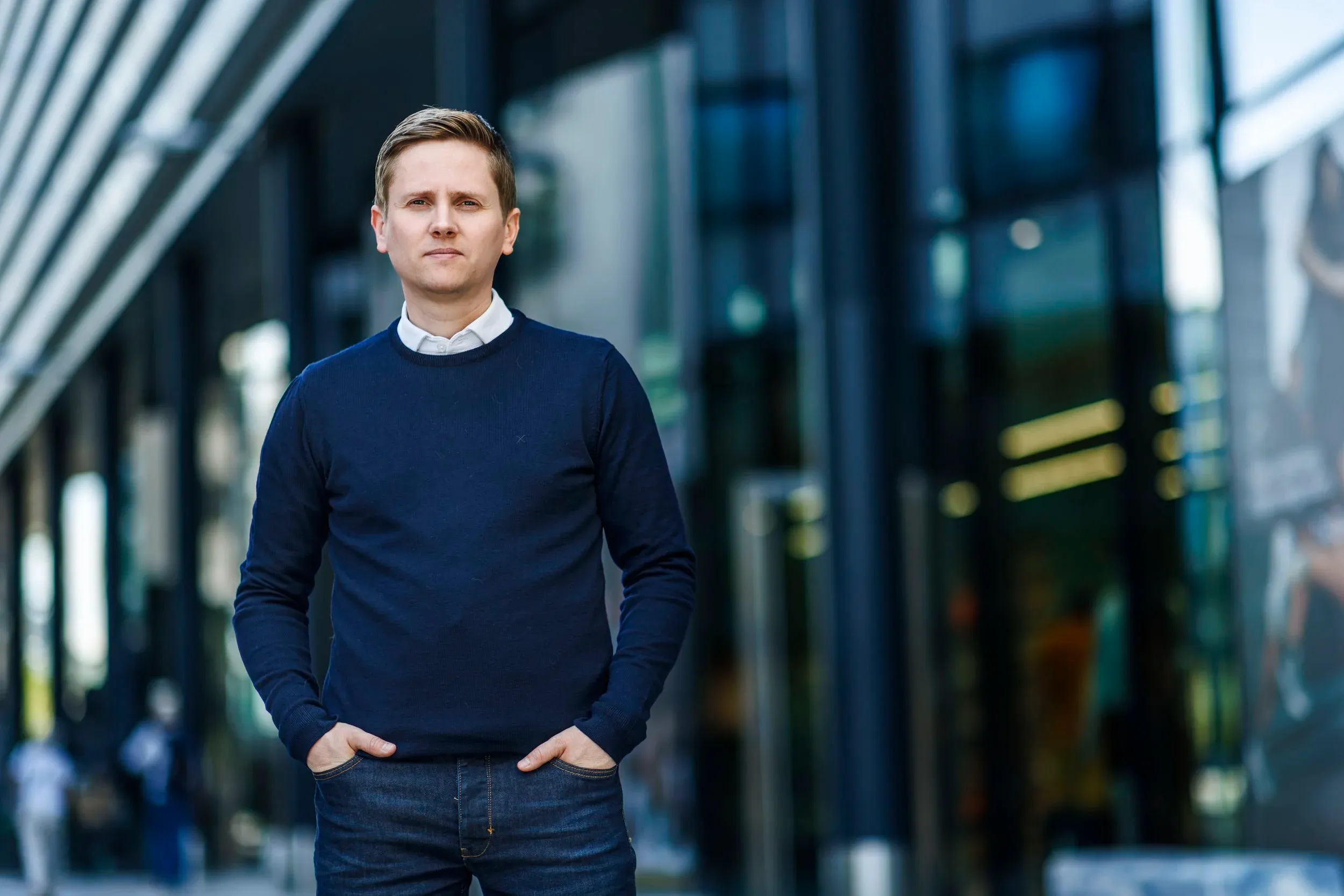 En ung mann i marineblå genser og jeans står med hendene i lommene foran en moderne glassbygning.
