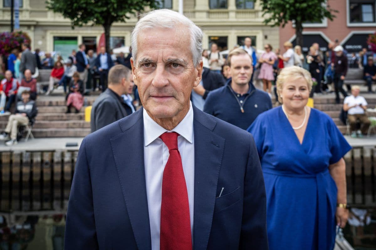 En eldre mann i dress og rødt slips står utendørs ved et vann, med folk samlet og sittende på trapper i bakgrunnen.