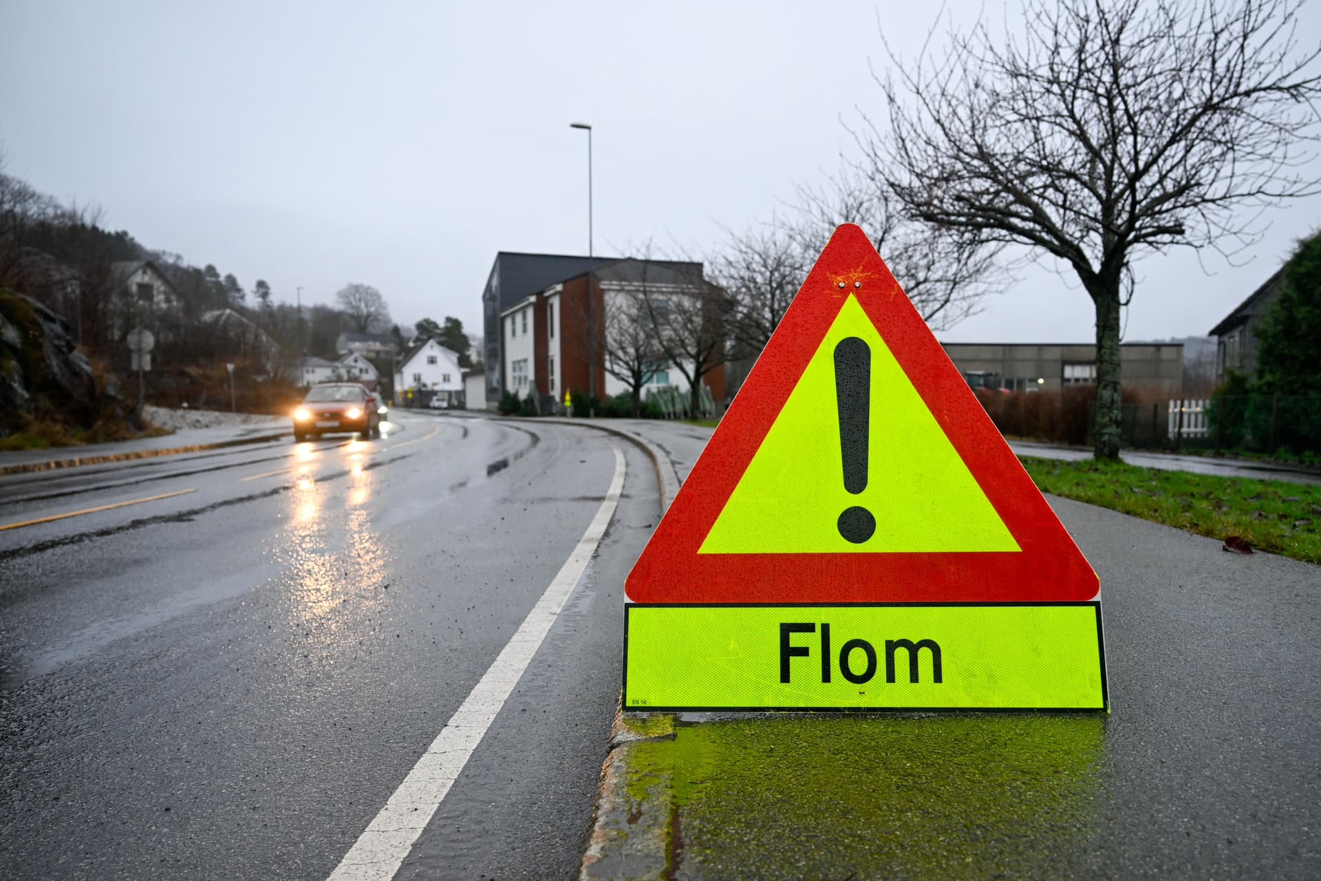 Et trekantet varselskilt med teksten «Flom» står ved en våt veikant, mens en bil nærmer seg i det fjerne.