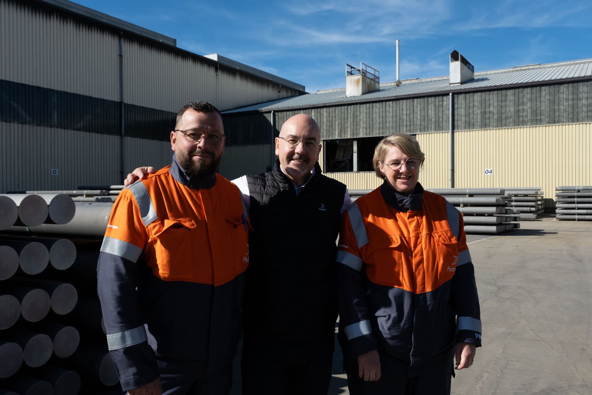 Tre personer, to i arbeidsjakker og én i vest, smiler utendørs foran en industribygning med stabler av metall.