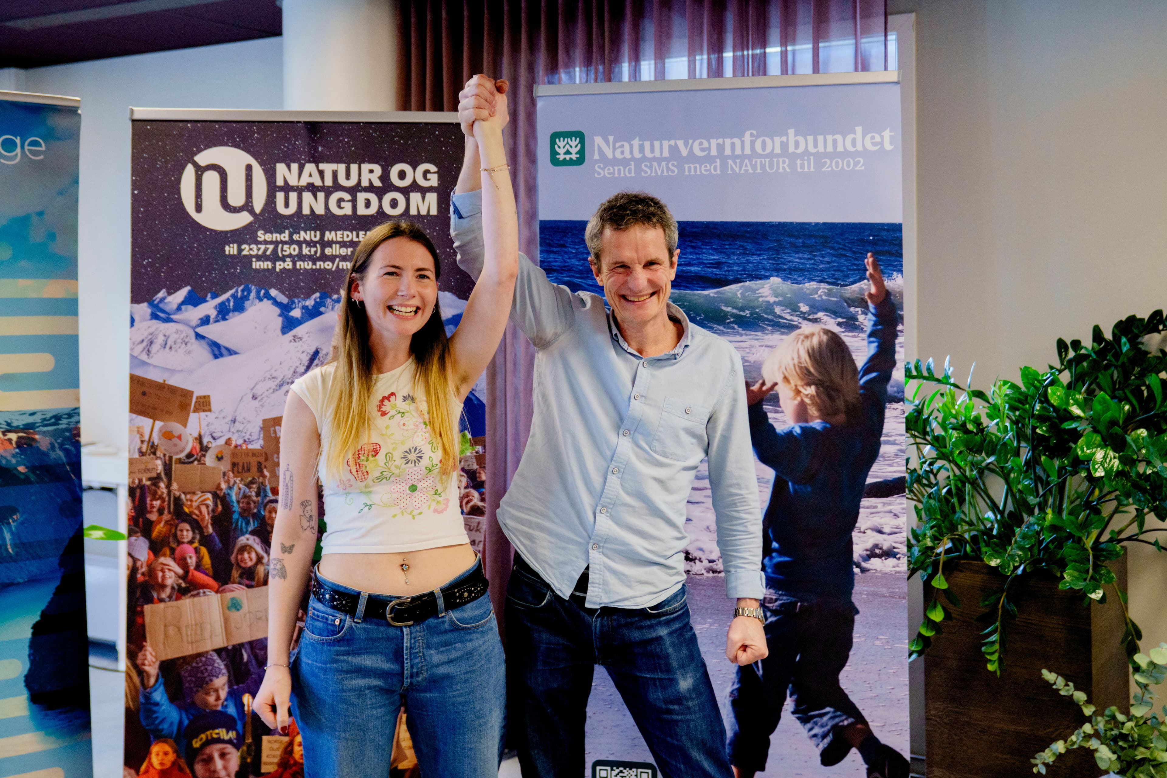 En smilende ung kvinne og mann løfter de sammenføyde hendene i triumf, flankert av bannere for «Natur og Ungdom» og «Naturvernforbundet».