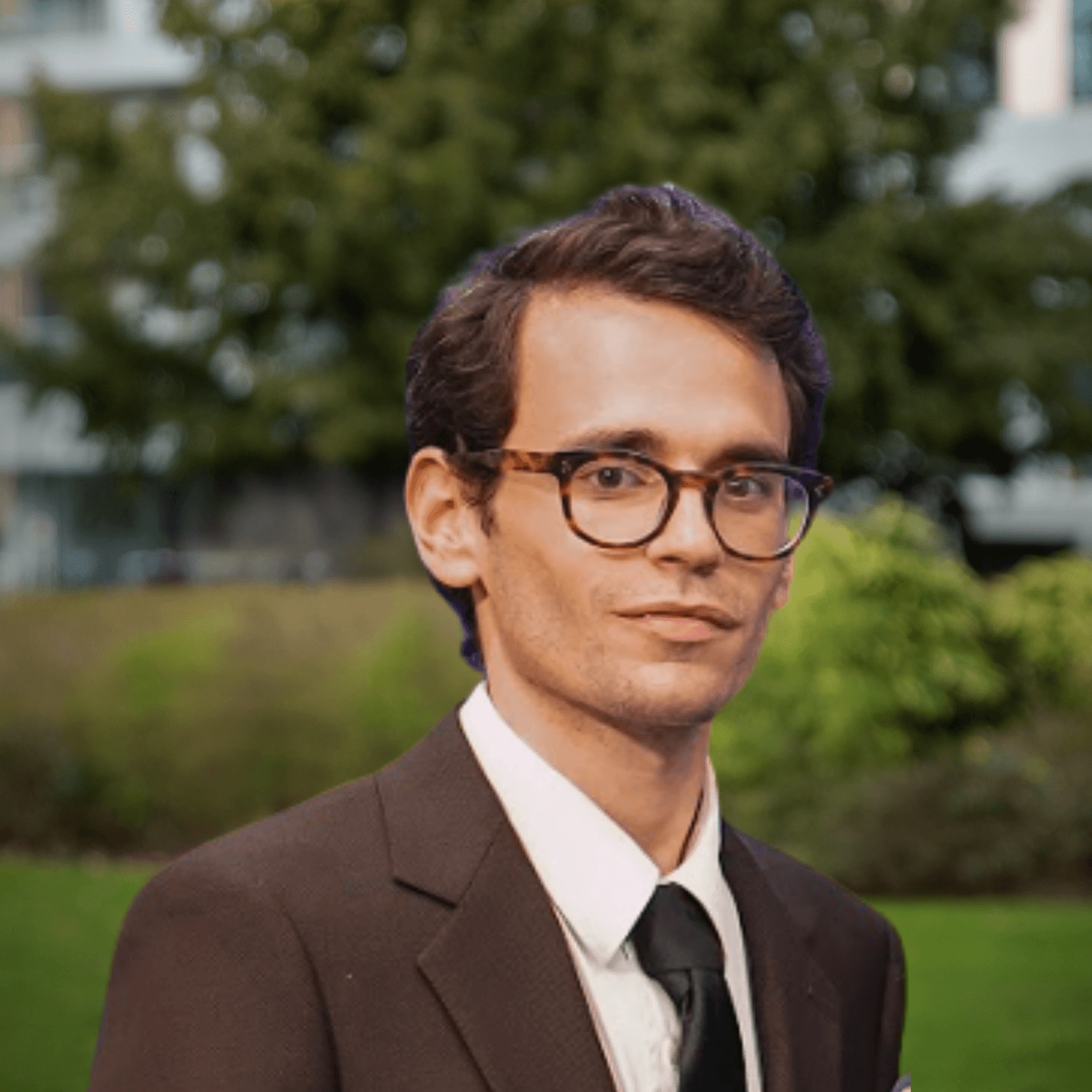 En mann med mørkt hår og skilpaddemønstrede briller, kledd i brun dress og svart slips, mot en uskarp grønn utendørs bakgrunn.