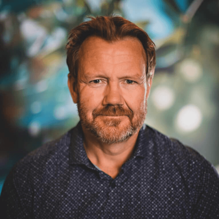 A man with reddish-brown hair and beard, wearing a blue patterned shirt, looks forward.