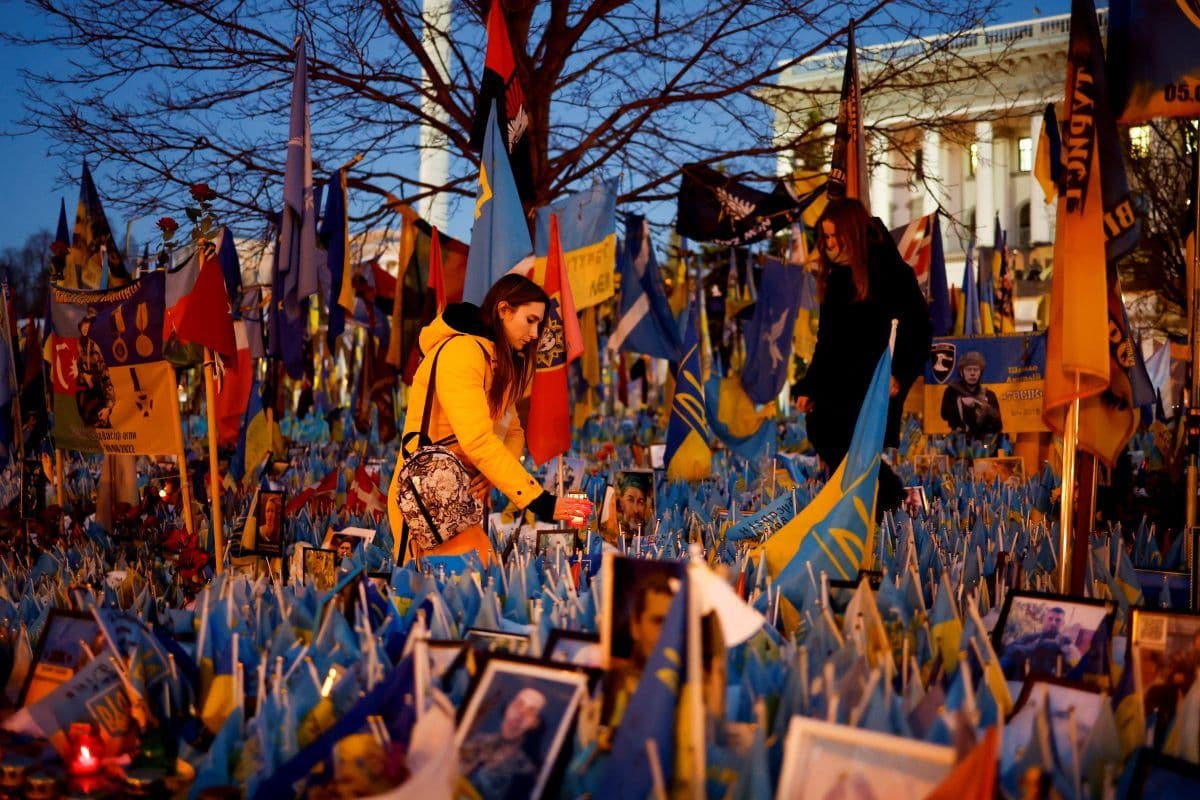 Folk arrangerer flagg og lys ved et minnesmerke med bilder utendørs, omgitt av mange blå og gule flagg, i en kveldssetting.
