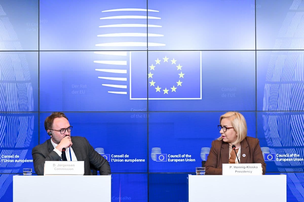 To tjenestemenn sitter ved et pressekonferansebord med navneskilt foran et blått bakteppe fra EU som viser EU-flagget og logoene til Rådet for Den europeiske union.