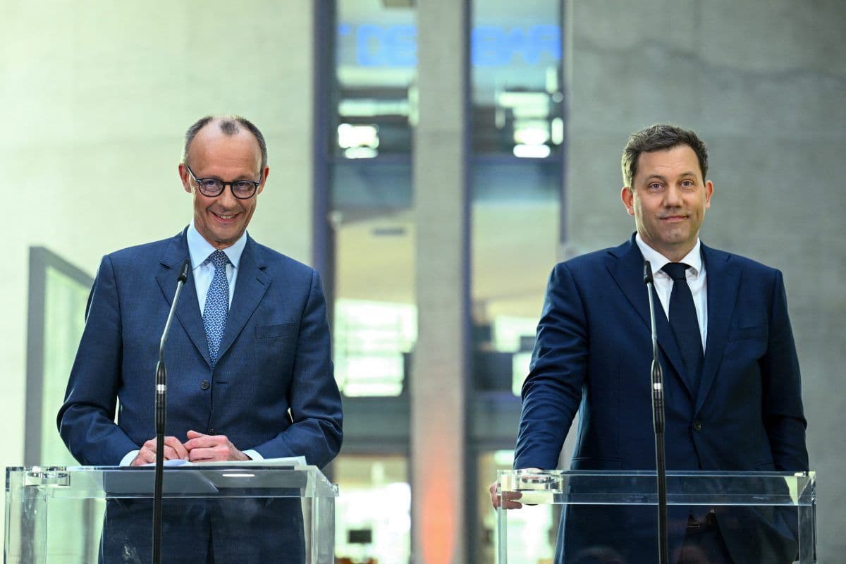 To menn i dress står ved podier innendørs, snakker i mikrofoner og gleder seg.