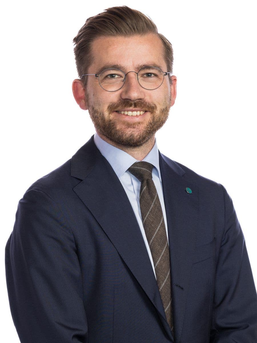 Smilende person med briller og skjegg, iført dress og slips, mot en hvit bakgrunn.