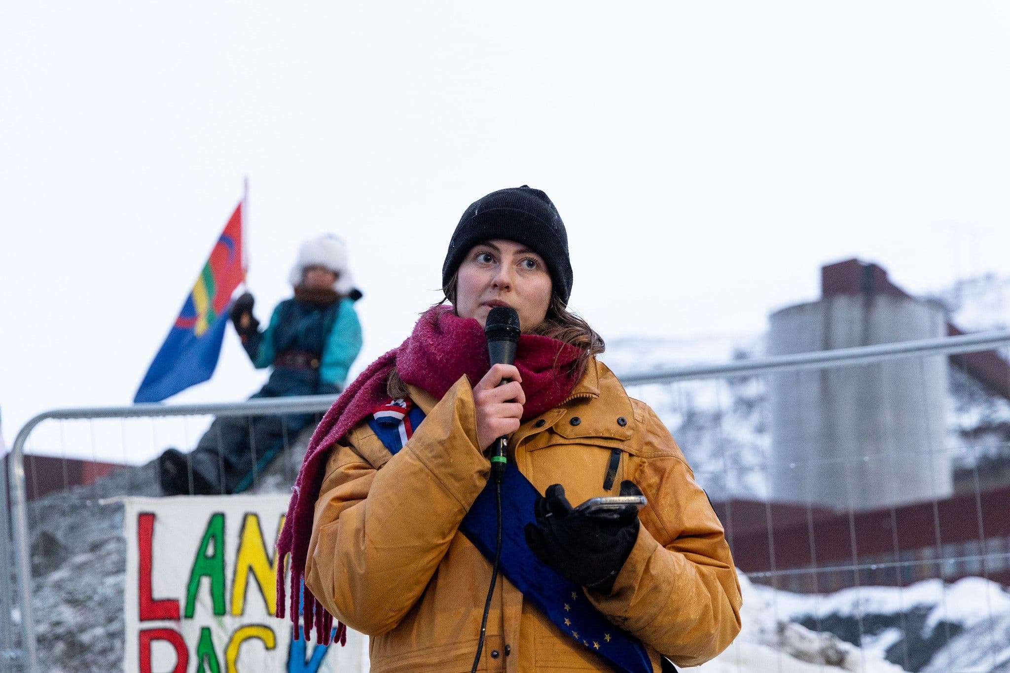 En kvinne i vinterklær og med et EU-bånd over skulderen snakker i en mikrofon, med et samisk flagg og et skilt med teksten «LAND BACK» synlig i den snødekte bakgrunnen.